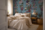 A bedroom interior featuring a bed dressed in beige linens, set against a prominent blue floral wallpaper with pink, purple, and green botanical patterns. A wooden nightstand with a pleated table lamp sits on the right, and a woven basket and wood floor are visible.