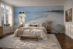 "Summer Evening on the Southern Beach" by Peder Severin Krøyer, made as a large wallpaper mural depicting a beach scene with two women in white dresses stands on the bedroom wall behind a bed with neutral bedding. The room has wood floors, a light rug, bedside tables, and a window on the left.
