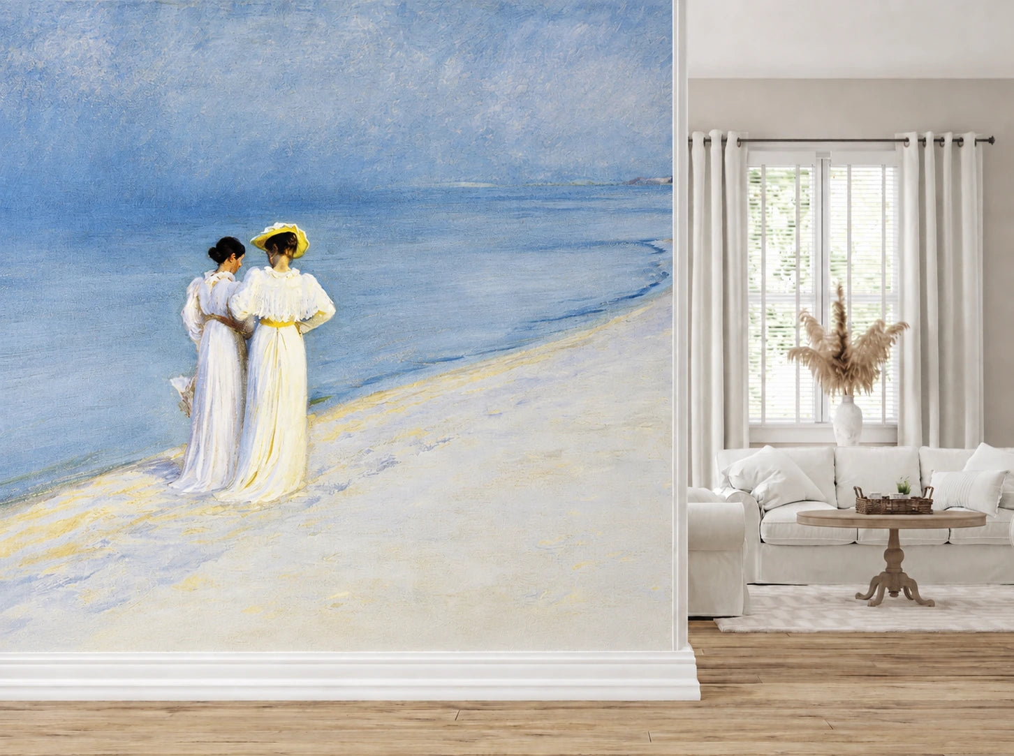 "Summer Evening on the Southern Beach" by Peder Severin Krøyer, made as a large wallpaper mural featuring an impressionist painting of two women in white dresses standing on a blue beach, shown installed in a modern living room next to a white sofa and window.