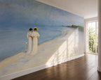 "Summer Evening on the Southern Beach" by Peder Severin Krøyer, made as a large wallpaper mural depicting a beach scene with two women in white dresses standing on a sandy beach under a blue sky, installed on a wall in a modern room with hardwood floors and a window.