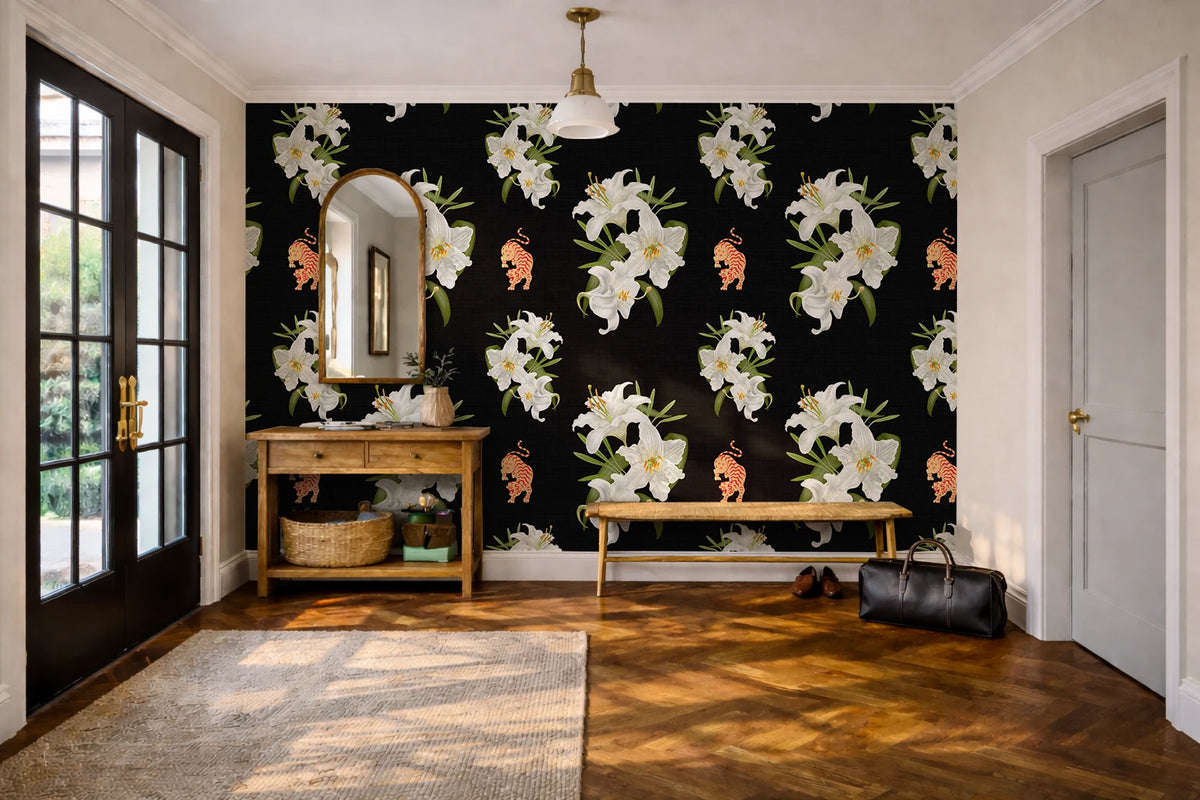 A styled interior room featuring a bold black wallpaper with large white lilies and stigers. The room includes a wooden console table with a mirror, a bench, a rug, wood flooring, and a black bag on the floor.