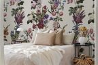 A styled bedroom featuring a wall covered in a vintage botanical floral wallpaper with pink, purple, and blue flowers on a cream background. The bed is dressed in cream-colored textured bedding with beige pillows, flanked by black side tables with a white lamp and a white vase.
