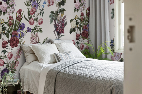 A styled bedroom featuring floral wallpaper with pink, purple, and green botanical patterns on a light background. A bed with white linens, a gray quilted coverlet, and decorative pillows is positioned against the wall, with gray curtains, a window, and green plants visible.