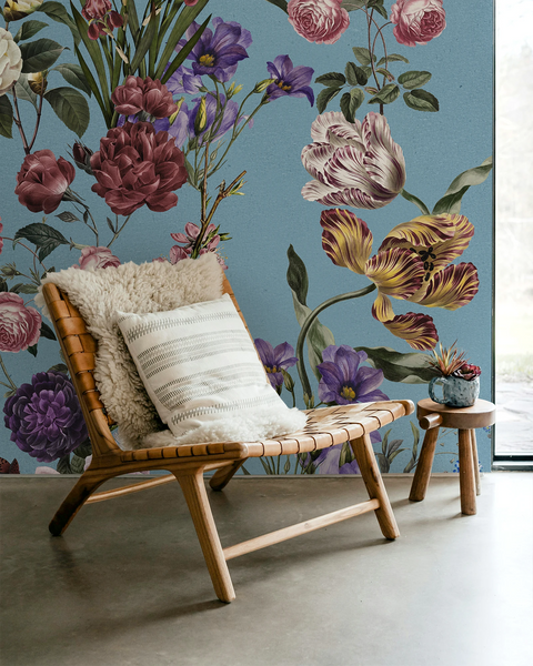 A styled interior scene featuring a woven wooden lounge chair with cream throw pillows, a small wooden stool, and a large floral wallpaper mural in muted blue, pink, purple, and yellow tones.