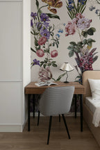 A styled bedroom corner featuring a large-scale botanical floral wallpaper mural with pink, purple, and green flowers. In front of the wall is a wooden writing desk with black legs, a black and white gingham patterned upholstered chair, a small black table lamp, and an open magazine.
