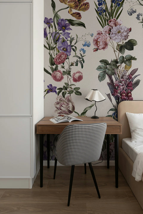 A styled bedroom corner featuring a large-scale botanical floral wallpaper mural with pink, purple, and green flowers. In front of the wall is a wooden writing desk with black legs, a black and white gingham patterned upholstered chair, a small black table lamp, and an open magazine.