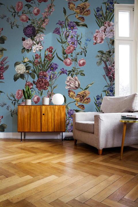 A styled interior room featuring a light blue wallpaper with a large multicolor floral pattern, paired with a light wood herringbone floor, a mid-century wooden cabinet, and a beige armchair.