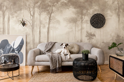 A styled living room featuring a large-scale forest tree wallpaper mural. A light gray sofa sits in the center with a white and black dog lying on it, surrounded by home decor including a black woven side table, a framed abstract print, and a small plant.