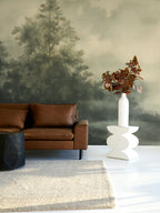 Modern living room with brown leather sofa, white vase with dried leaves, and a moody forest wallpaper mural. 