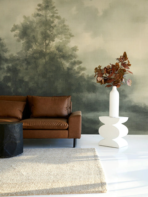 Modern living room with brown leather sofa, white vase with dried leaves, and a moody forest wallpaper mural. 