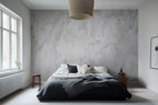 A bedroom with the Roma Cloud Marble wallpaper covering the wall behind the bed, featuring a creamy marble-like pattern with soft grey swirls.