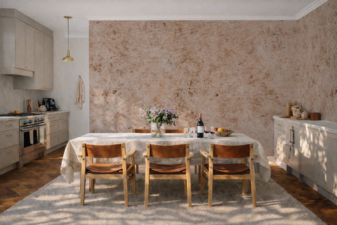 A styled dining room scene featuring a industrial textured beige wallpaper accent wall, a light wood dining table with a white tablecloth and four wooden chairs with brown leather seats, set on a light beige rug.