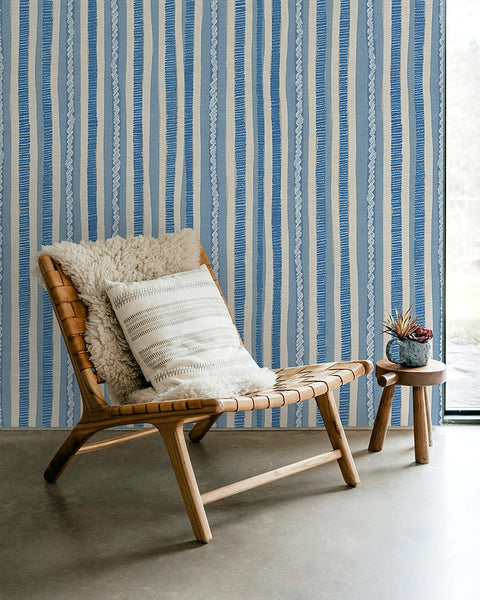 Wooden chair with patterned cushions against a blue and white striped wallpaper.