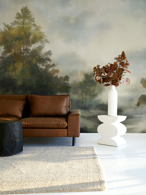 Modern living room with brown leather sofa, white side table, and nature-themed wallpaper mural.