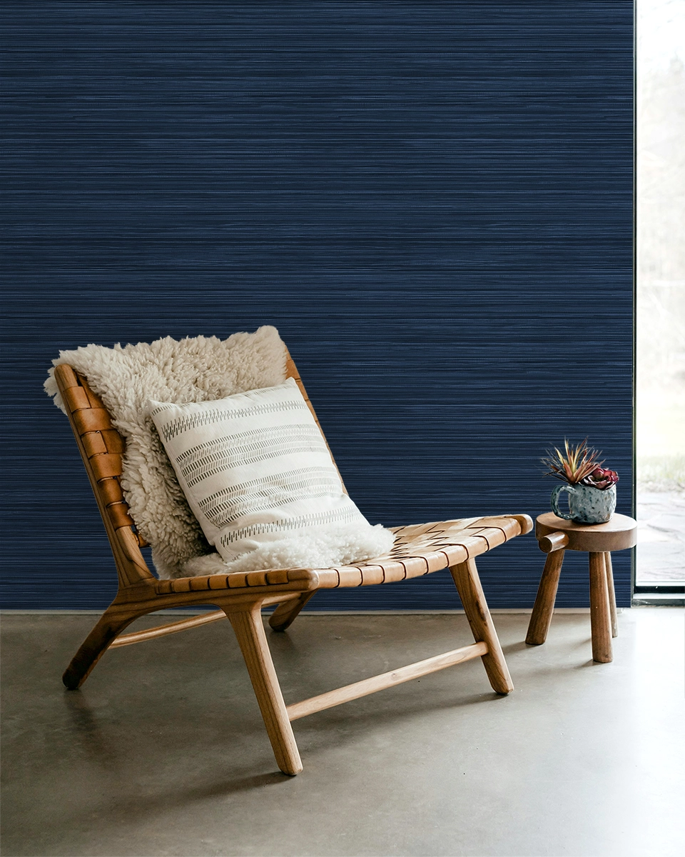 Navy blue faux grasscloth wallpaper with a rich woven texture, in a room with a lounge chair and a side table with flowers.
