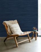 Navy blue faux grasscloth wallpaper with a rich woven texture, in a room with a lounge chair and a side table with flowers.
