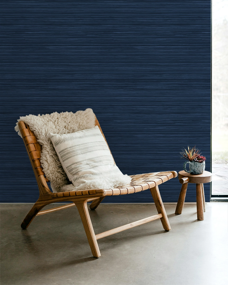 Navy blue faux grasscloth wallpaper with a rich woven texture, in a room with a lounge chair and a side table with flowers.

