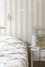Bedroom with striped wallpaper, bed with white bedding, and nightstand with lamp.