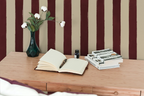 Open book on a wooden desk with a vase and stationery items against a wall with striped wallpaper.