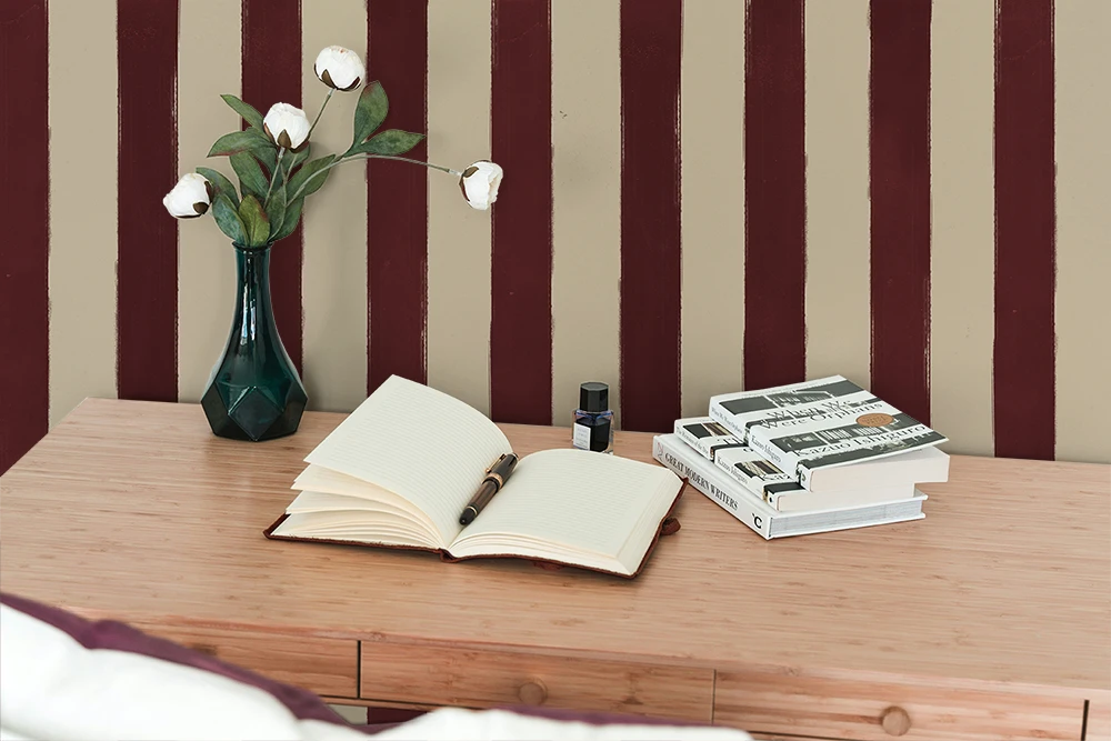 Open book on a wooden desk with a vase and stationery items against a wall with striped wallpaper.