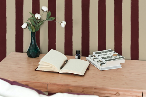 Open book on a wooden desk with a vase and stationery items against a wall with striped wallpaper.
