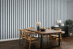 A styled dining room featuring a prominent wall of vertical navy blue and white striped wallpaper. In the foreground, a rustic wooden dining table with black metal legs is surrounded by four wooden chairs.