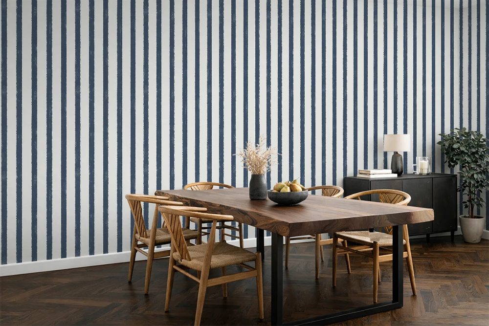 A styled dining room featuring a prominent wall of vertical navy blue and white striped wallpaper. In the foreground, a rustic wooden dining table with black metal legs is surrounded by four wooden chairs.