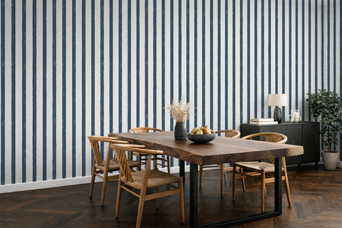 A styled dining room featuring a prominent wall of vertical navy blue and white striped wallpaper. In the foreground, a rustic wooden dining table with black metal legs is surrounded by four wooden chairs.