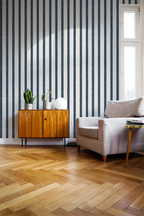 A styled interior room with vertical navy and white striped wallpaper, light oak herringbone wood flooring, a mid-century wooden sideboard with potted plants, a light gray armchair, and a small round side table.