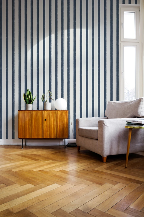 A styled interior room with vertical navy and white striped wallpaper, light oak herringbone wood flooring, a mid-century wooden sideboard with potted plants, a light gray armchair, and a small round side table.