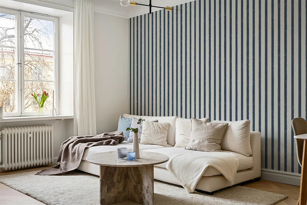 A styled living room corner featuring a cream-colored sectional sofa with neutral throw pillows, a round stone coffee table, and a prominent blue and white vertically striped wallpaper on the accent wall.