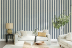 A styled living room featuring a bold navy and white vertical stripe wallpaper as the primary product. In front of the wall is a white sofa with neutral and mustard yellow pillows, a wooden coffee table, a side table with a lamp, and a tall potted olive tree.