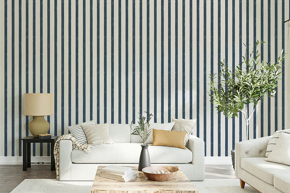 A styled living room featuring a bold navy and white vertical stripe wallpaper as the primary product. In front of the wall is a white sofa with neutral and mustard yellow pillows, a wooden coffee table, a side table with a lamp, and a tall potted olive tree.