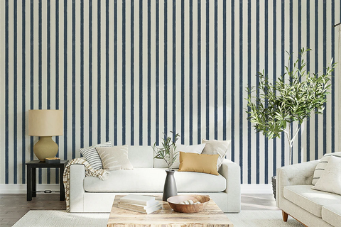 A styled living room featuring a bold navy and white vertical stripe wallpaper as the primary product. In front of the wall is a white sofa with neutral and mustard yellow pillows, a wooden coffee table, a side table with a lamp, and a tall potted olive tree.