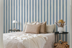 A styled bedroom scene with a bed dressed in cream bedding and multiple beige and gray pillows, set against a blue-and-white vertically striped wall. Two black side tables flank the bed, one with a white table lamp and the other with a white vase of dried flowers; a woven basket bag sits on the floor nearby.