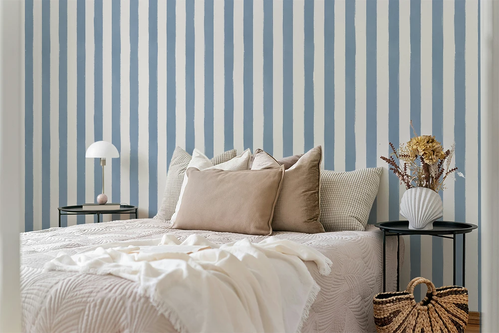 A styled bedroom scene with a bed dressed in cream bedding and multiple beige and gray pillows, set against a blue-and-white vertically striped wall. Two black side tables flank the bed, one with a white table lamp and the other with a white vase of dried flowers; a woven basket bag sits on the floor nearby.