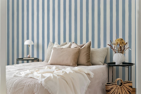 A styled bedroom scene with a bed dressed in cream bedding and multiple beige and gray pillows, set against a blue-and-white vertically striped wall. Two black side tables flank the bed, one with a white table lamp and the other with a white vase of dried flowers; a woven basket bag sits on the floor nearby.