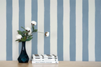Blue and white vertically striped wallpaper shown on a wall behind a wooden table with a dark blue vase holding white flowers and a stack of books.