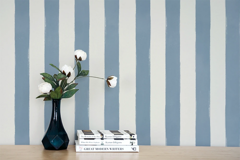 Blue and white vertically striped wallpaper shown on a wall behind a wooden table with a dark blue vase holding white flowers and a stack of books.