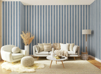 A styled living room featuring a prominent wall of vertical blue and white wallpaper stripes, with a white sofa, cream armchair, round ottoman, and a small round coffee table on a woven rug.