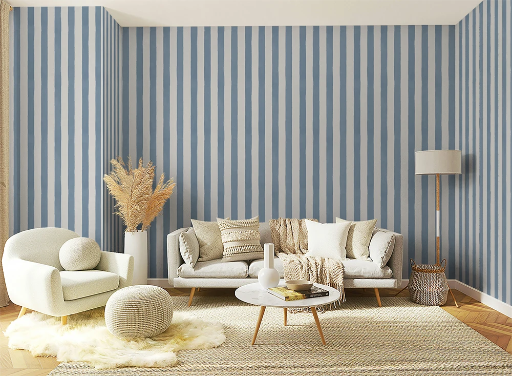 A styled living room featuring a prominent wall of vertical blue and white wallpaper stripes, with a white sofa, cream armchair, round ottoman, and a small round coffee table on a woven rug.
