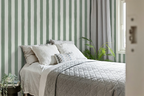A styled bedroom scene featuring a bed with white bedding and a gray quilted coverlet, set against a wall with wallpaper in vertical sage green and white stripes. Gray curtains hang by a window, and a green houseplant sits on the floor.