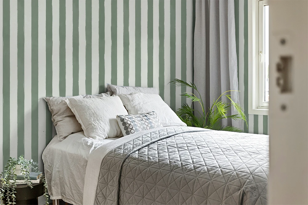 A styled bedroom scene featuring a bed with white bedding and a gray quilted coverlet, set against a wall with wallpaper in vertical sage green and white stripes. Gray curtains hang by a window, and a green houseplant sits on the floor.