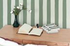 A styled interior scene showing a wooden desk against a green and white striped wallpaper. On the desk are an open notebook with a pen, a small dark bottle, and a stack of books, alongside a dark green vase holding white cotton flowers.