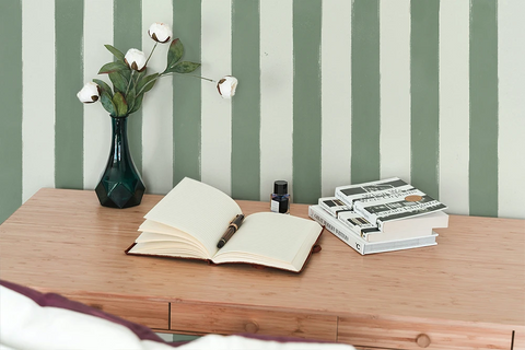 A styled interior scene showing a wooden desk against a green and white striped wallpaper. On the desk are an open notebook with a pen, a small dark bottle, and a stack of books, alongside a dark green vase holding white cotton flowers.