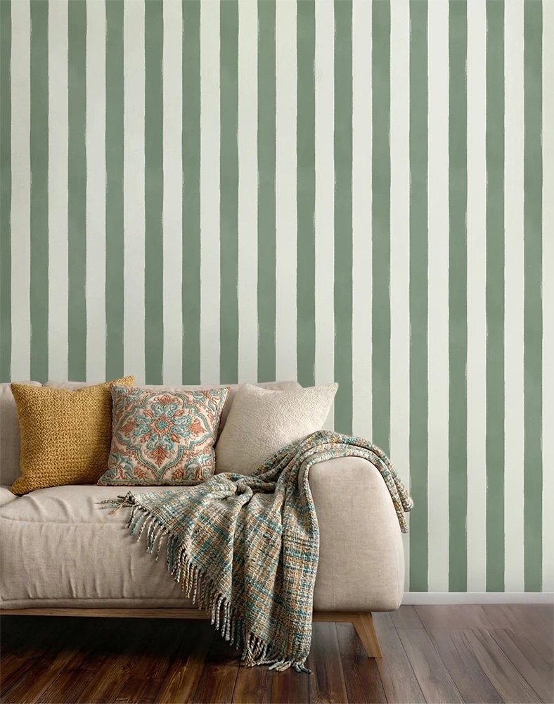 A styled living room corner featuring a beige sofa with decorative pillows and a fringed throw blanket, set against a wall with vertical sage green and off-white striped wallpaper and dark wood flooring.