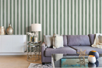 A styled living room featuring a lavender-gray fabric sofa with neutral throw pillows, set against a wallpaper of vertical sage green and white stripes. A white sideboard with decorative vases, a small round glass side table with a lamp, and a glass-top coffee table with candles are arranged in the foreground.