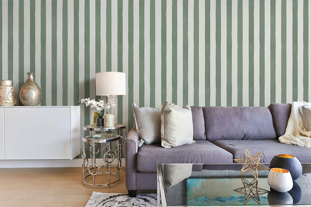 A styled living room featuring a lavender-gray fabric sofa with neutral throw pillows, set against a wallpaper of vertical sage green and white stripes. A white sideboard with decorative vases, a small round glass side table with a lamp, and a glass-top coffee table with candles are arranged in the foreground.