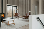 A modern living room interior with vertical brown and beige striped wallpaper, a black wood-burning stove, two brown leather armchairs, a white coffee table, a light patterned rug, and a white staircase railing in the foreground.