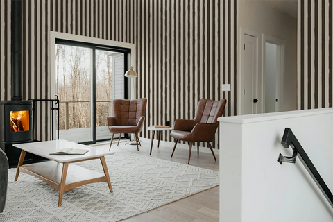 A modern living room interior with vertical brown and beige striped wallpaper, a black wood-burning stove, two brown leather armchairs, a white coffee table, a light patterned rug, and a white staircase railing in the foreground.
